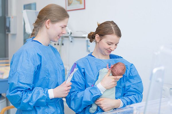 Das Bild zeigt zwei Pflegekräfte, von denen eine ein Neugeborenes auf dem Arm hat