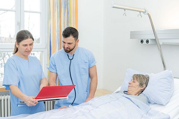 Das Bild zeigt zwei Fachkräfte bei der Pflegevisite am Bett einer Patientin